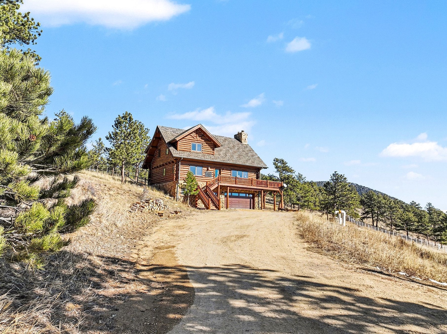 Rustic log cabin nestled on a hillside surrounded by pine trees and rolling mountain terrain under bright blue skies.