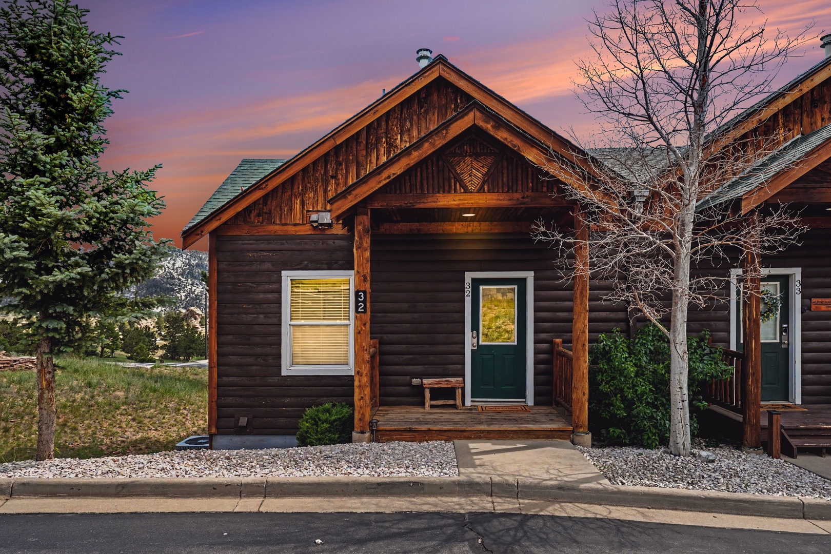 Rustic cabin exterior with warm wood siding, welcoming front porch, and mountain-style architecture, set against a stunning sunset sky with vibrant pink and orange hues.