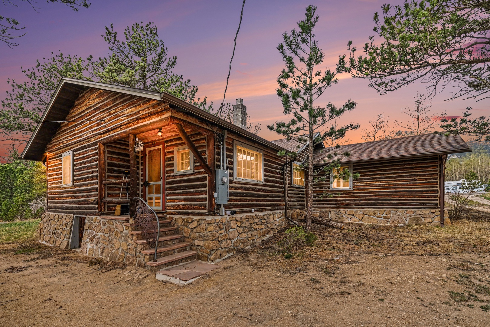 Rustic log cabin with stone foundation nestled among evergreen and aspen  trees, photographed during twilight hours with dramatic purple-orange sky.