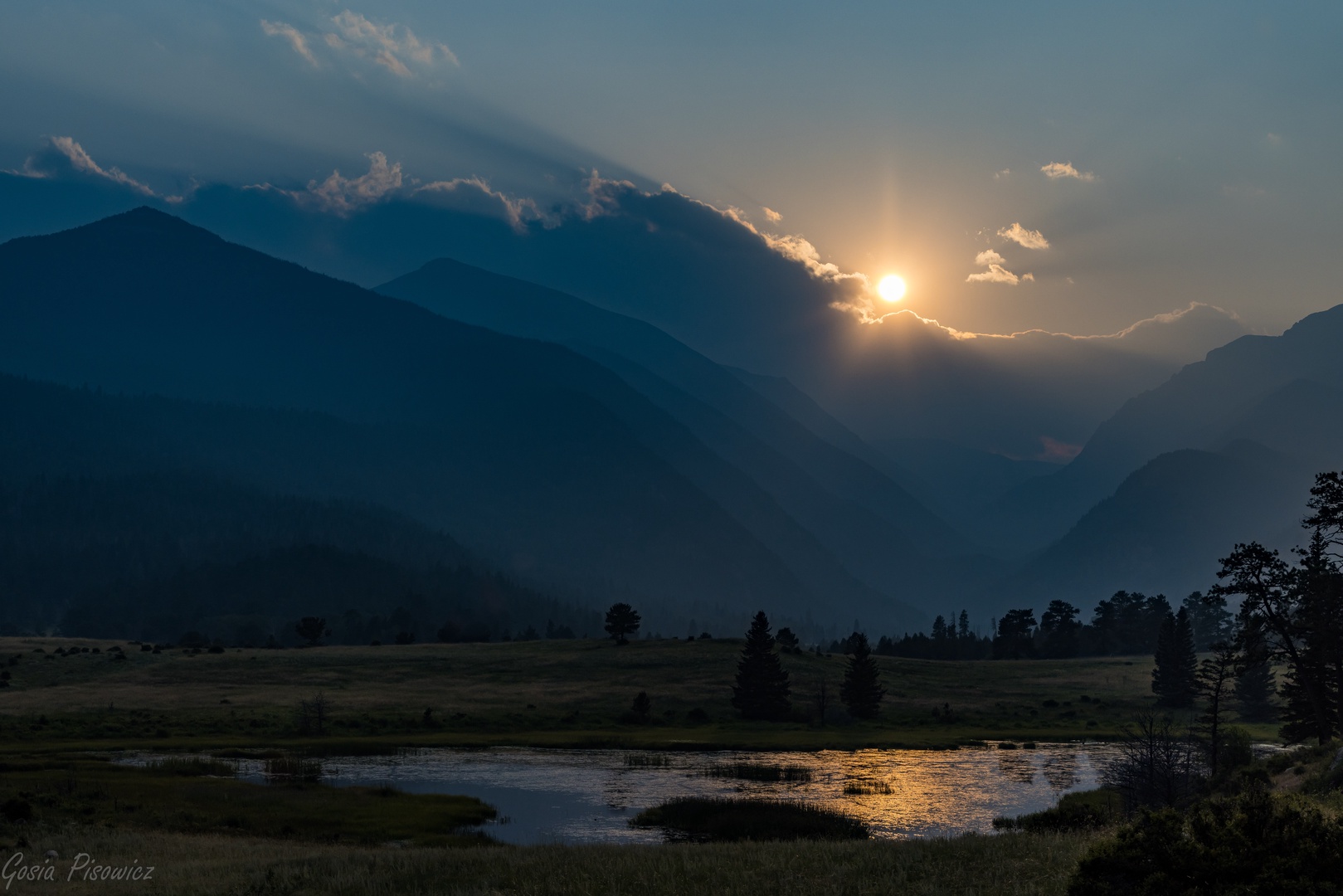 A stunning mountain landscape surrounding the property features dramatic peaks, open meadows, and a tranquil water feature as the sun sets behind layered ridges.