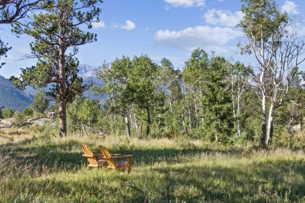 Two wooden Adirondack chairs sit in a peaceful meadow surrounded by aspens and mountain views, offering a perfect spot to unwind in nature.