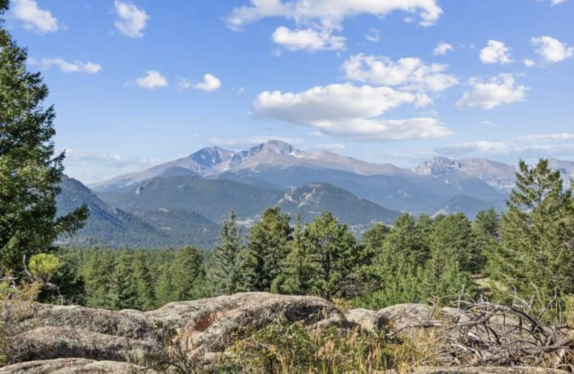 Majestic Longs Peak and the Front Range rise above forested foothills under a bright blue sky dotted with white clouds.