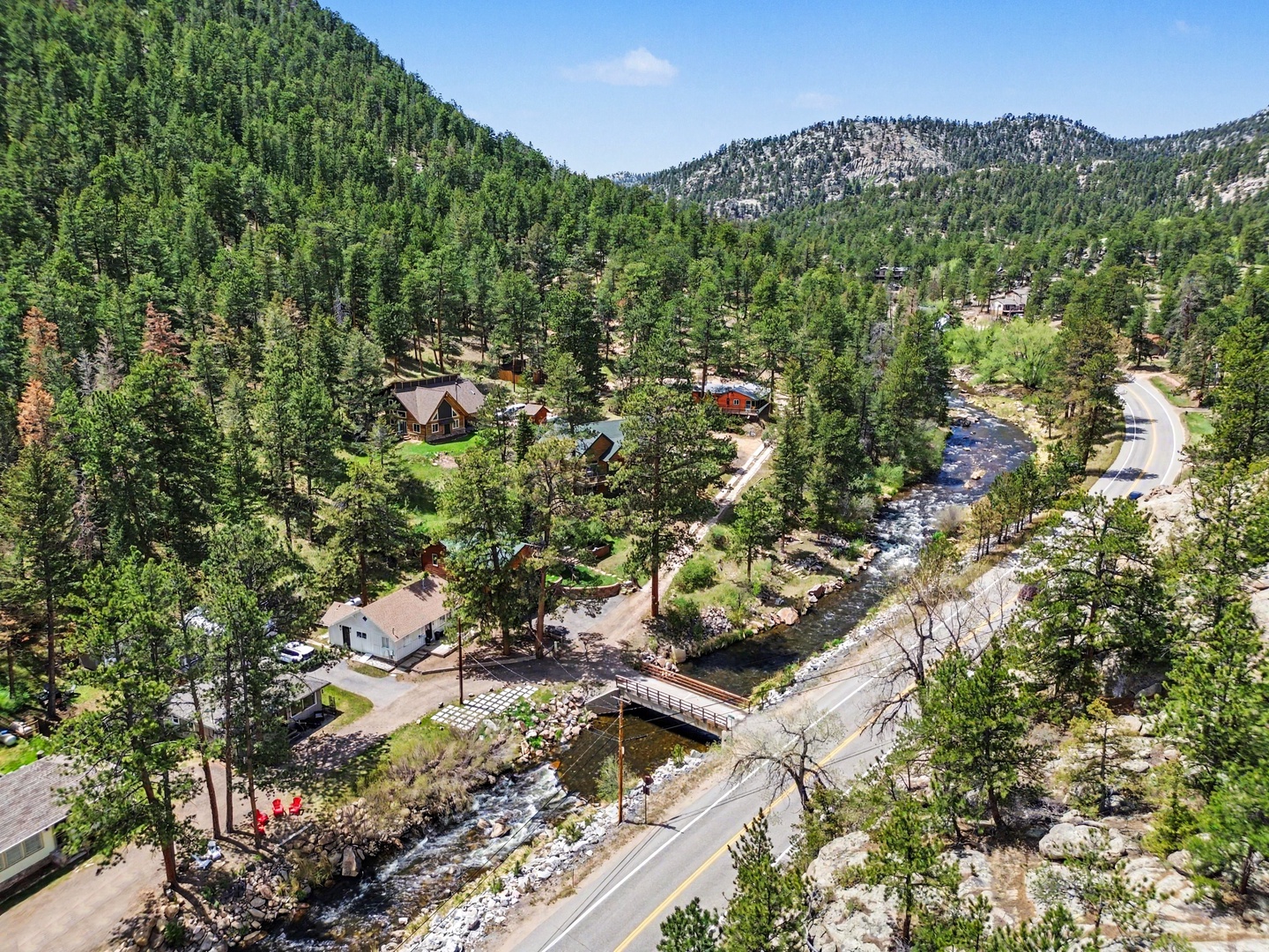 An aerial view reveals a peaceful mountain retreat nestled along a flowing river, surrounded by pine forests and scenic mountain peaks in a quiet neighborhood.