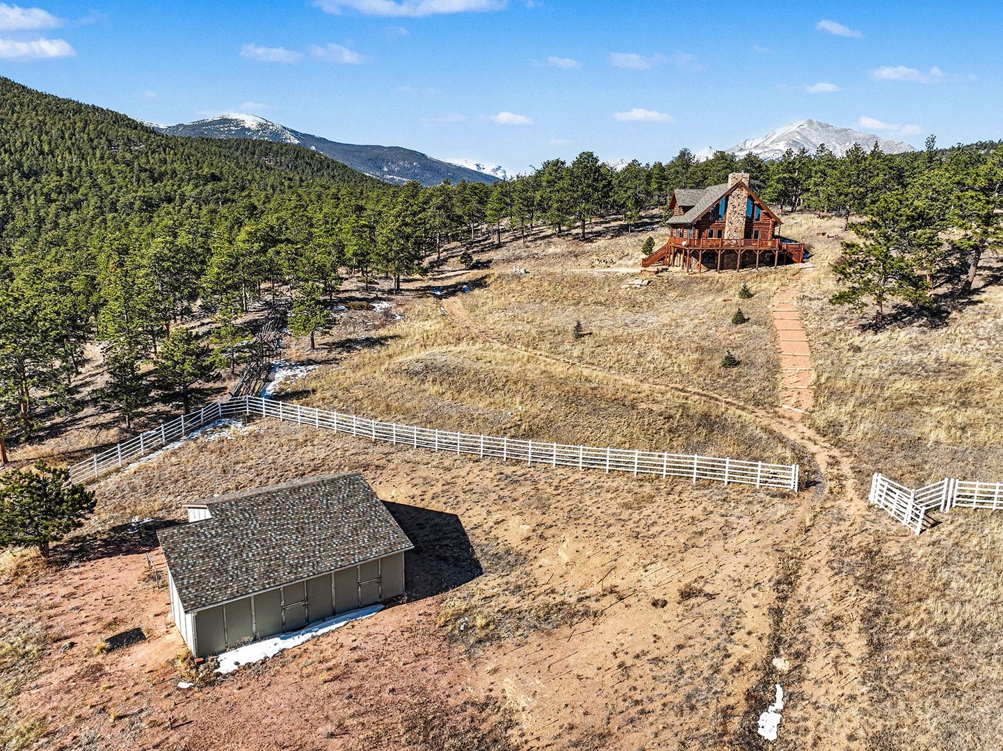 Aerial view of mountain property featuring rustic cabin and outbuildings surrounded by pristine wilderness and snow-capped peaks.