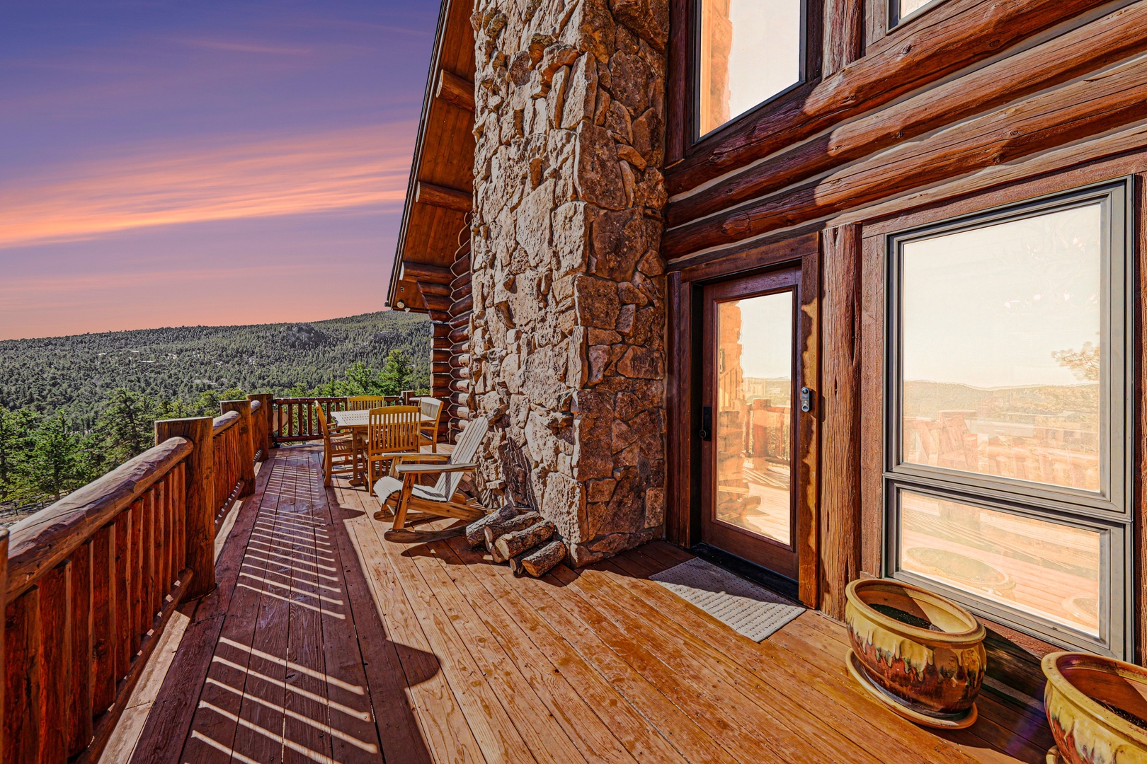 Rustic log cabin with stone accents and wraparound deck overlooking forested mountains at sunset.
