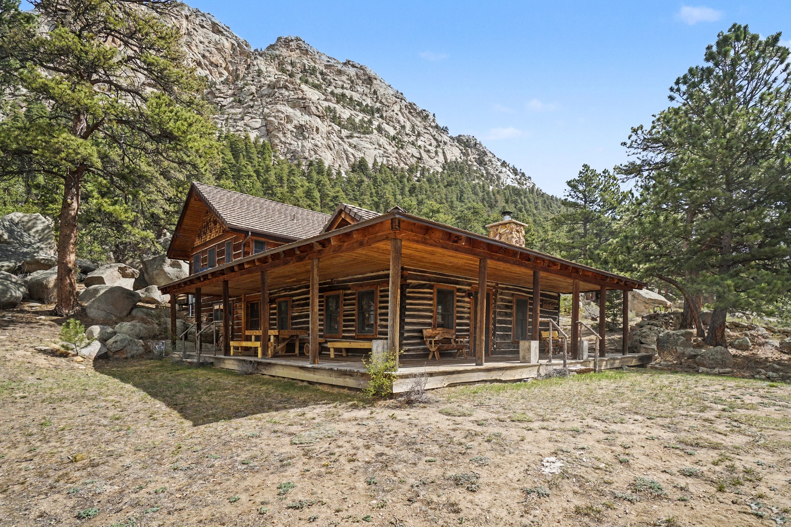 A stunning log cabin nestled beneath dramatic mountain cliffs, surrounded by towering pines and natural boulder formations.