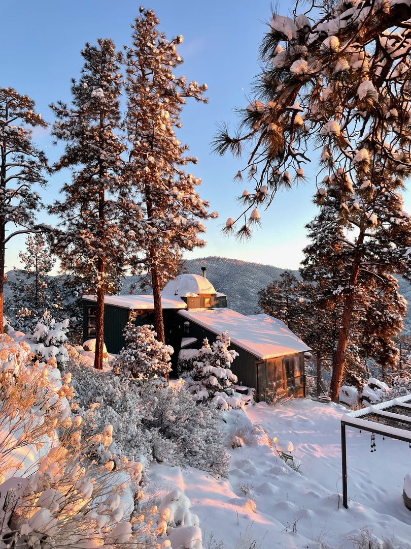 A secluded mountain retreat nestled among snow-frosted pines, where golden sunset light bathes the property in winter's serene embrace—part of the beautiful surrounding landscape.