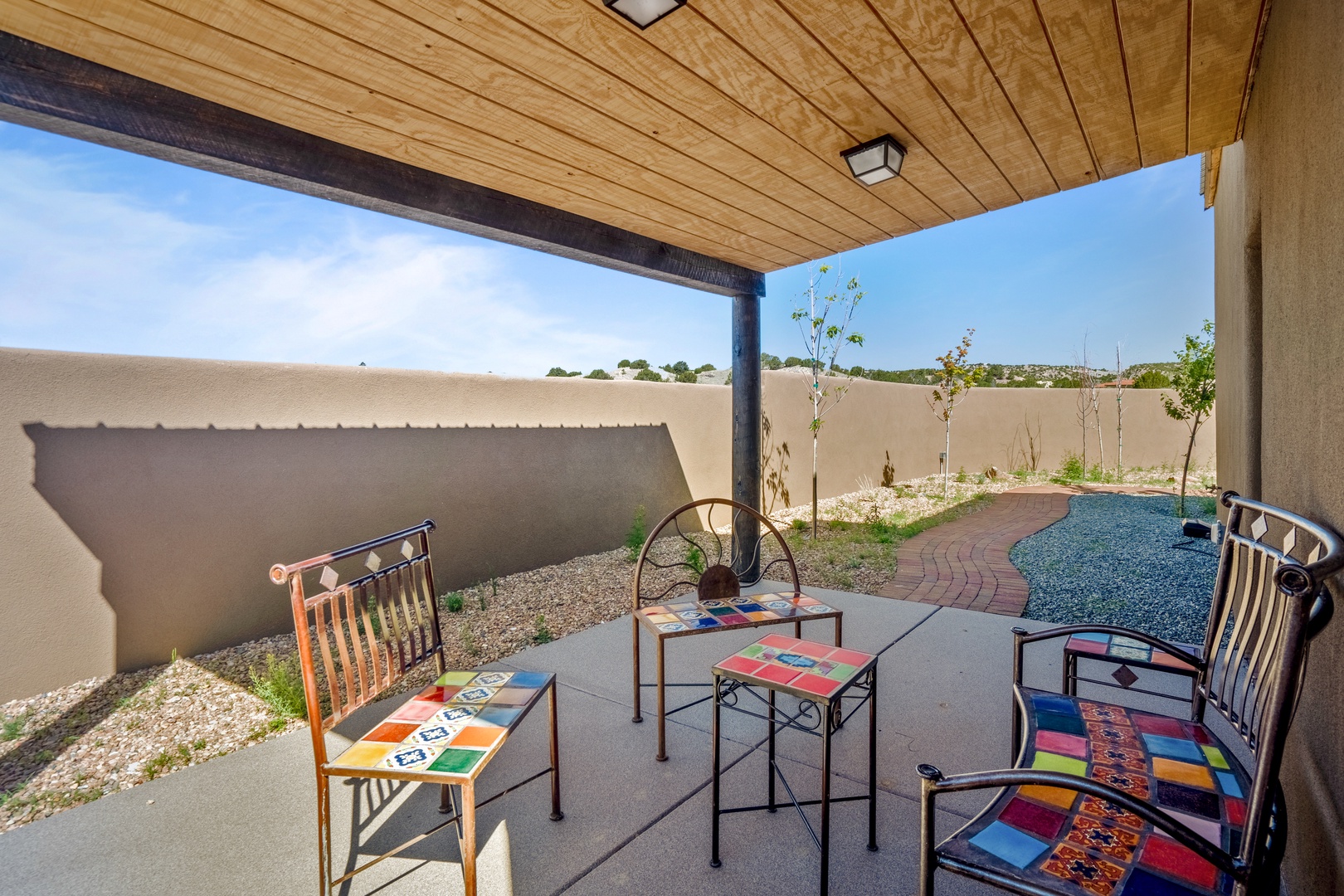 Step onto your private covered patio where colorful seating awaits under warm wooden beams, overlooking your peaceful garden courtyard.