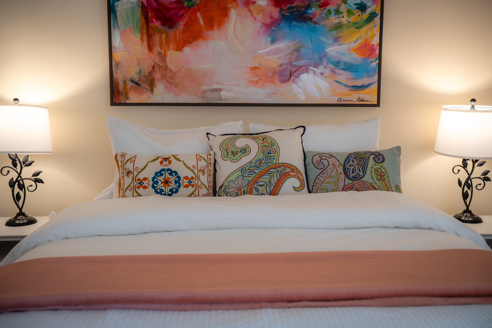 Settle into your comfortable bed adorned with vibrant paisley pillows, where soft lighting and colorful artwork create a welcoming retreat for your stay.