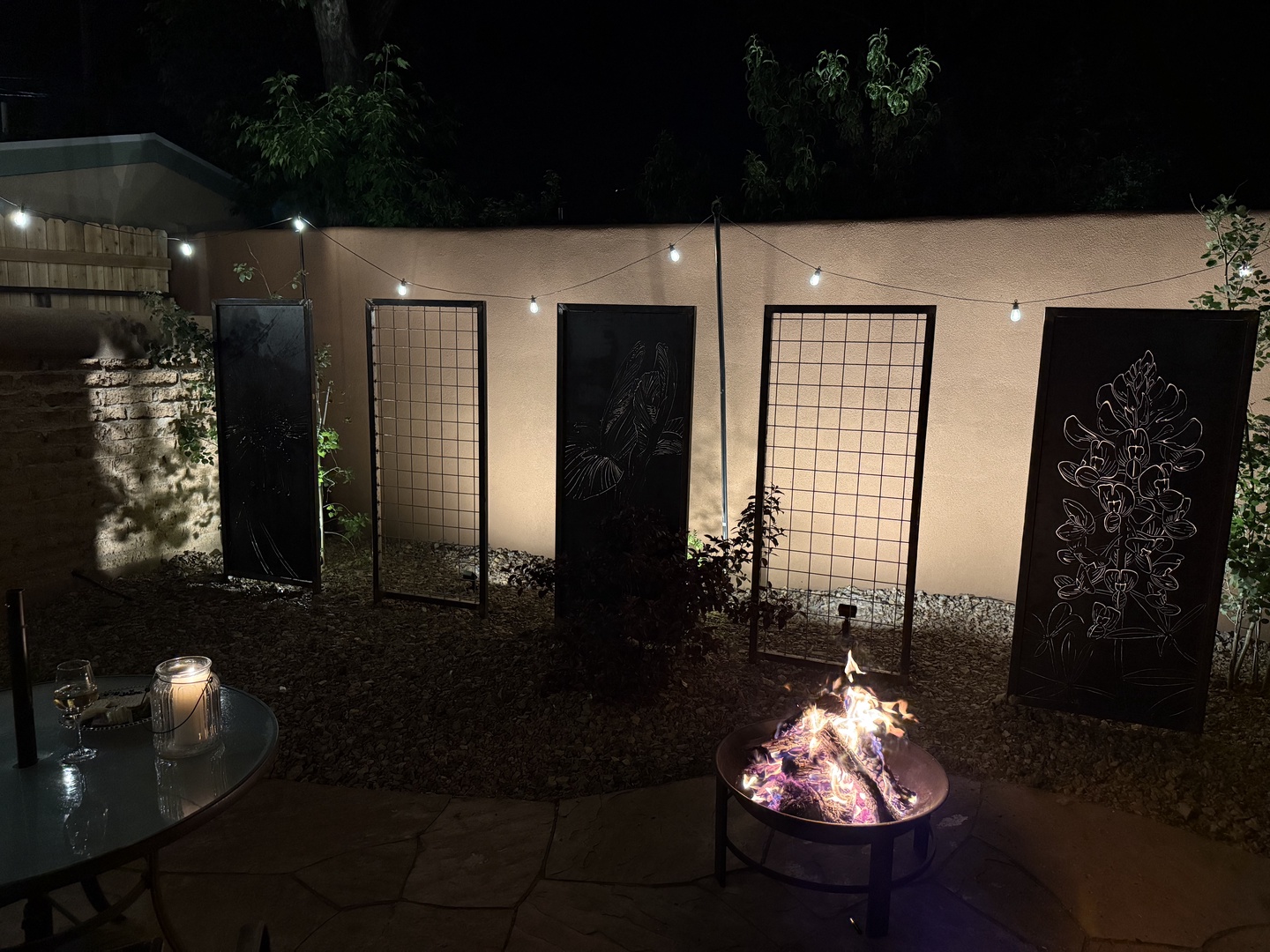 Gather around the glowing fire pit in your private outdoor courtyard, where string lights cast a warm glow over the artistic panels and intimate seating—perfect for evening conversations under the stars.
