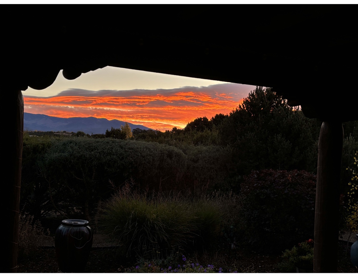 A dramatic sunset paints the sky in brilliant orange hues, framed by the property's entrance and surrounded by lush natural landscape and distant mountain ranges.