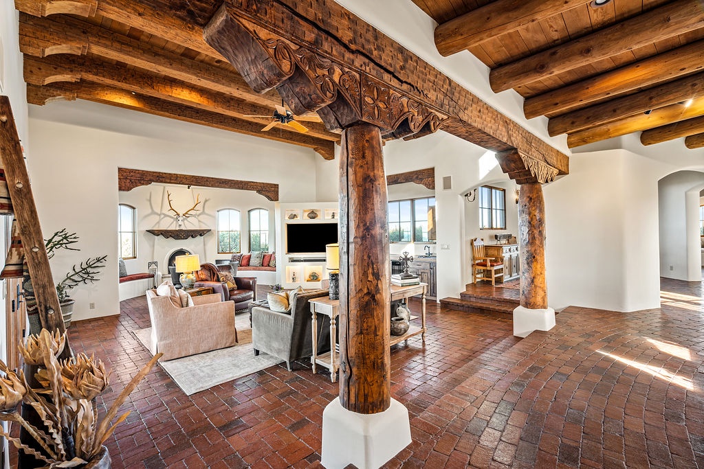 Step into your grand southwestern retreat where handcrafted vigas frame soaring ceilings and warm brick floors ground this stunning open-concept living space.