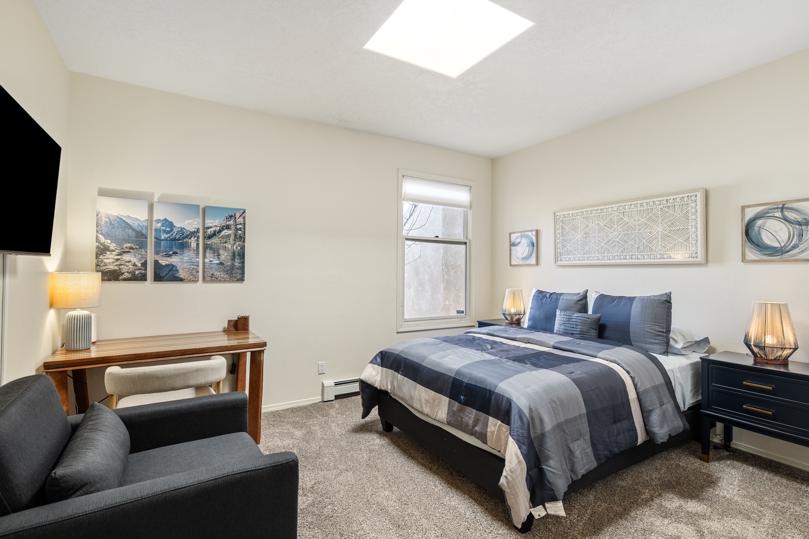 Your comfortable bedroom features a cozy queen bed with plaid bedding, convenient workspace, armchair for relaxing, and natural light from the skylight above.