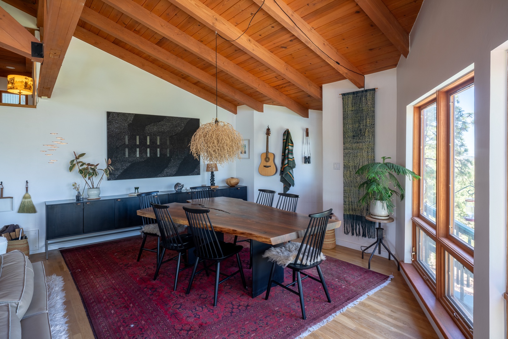 Gather around this stunning handcrafted dining table beneath a tumbleweed light and soaring wooden beams. Natural light pours through floor-to-ceiling windows, illuminating artisan touches and cozy textures throughout this inviting space.