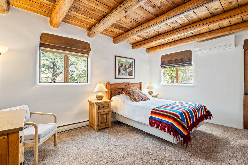 Unwind in your cozy adobe-style bedroom featuring rustic vigas overhead, colorful southwestern textiles, and peaceful garden views through dual windows.