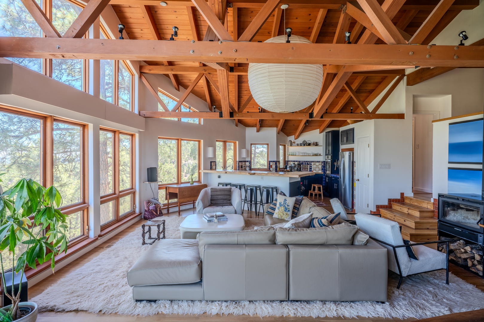 Relax into this airy living space where exposed beams frame forest views through floor-to-ceiling windows. You'll love lounging on the spacious sofa as sunlight pours in, connecting you to nature without leaving your cozy retreat.