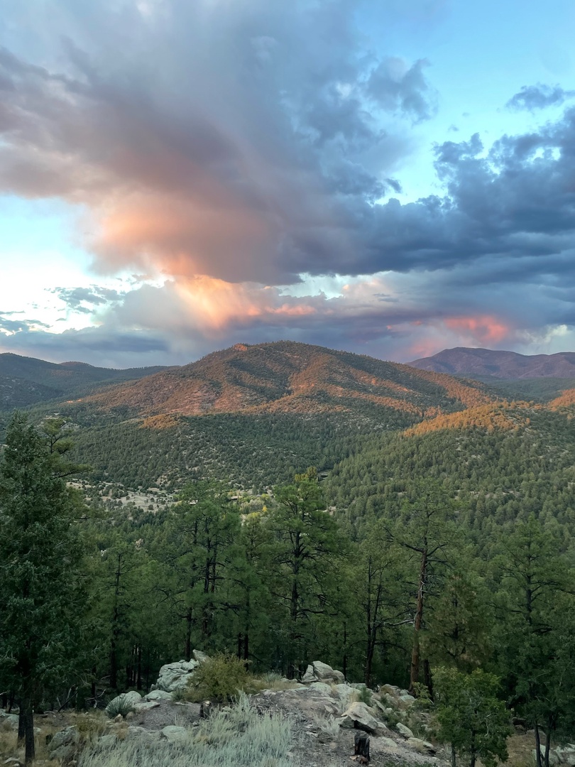 A dramatic mountain landscape surrounds the property, with forested ridges and valleys stretching into the distance beneath a colorful sky at day's end.