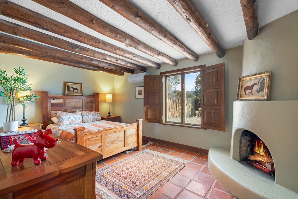 Cozy up in your charming bedroom with rustic wooden beams, crackling fireplace, and peaceful garden views through traditional shutters.