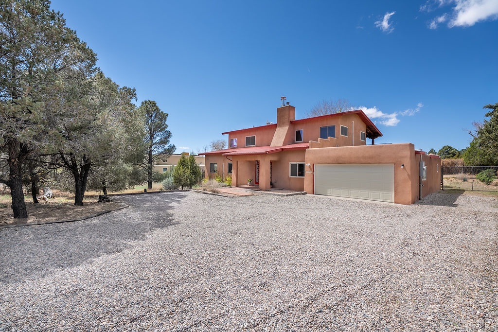 Charming adobe-style home with spacious gravel driveway and attached garage, nestled among mature trees under brilliant blue skies.