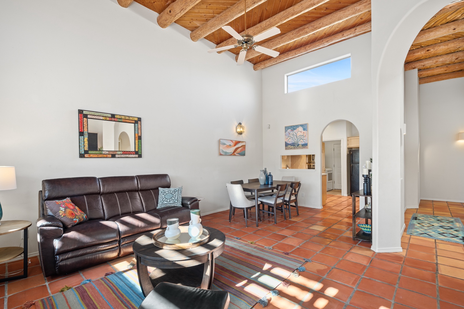 Relax in your open-plan living space featuring rustic wooden ceiling beams, comfortable leather seating, and warm terra cotta floors that create an inviting southwestern atmosphere.