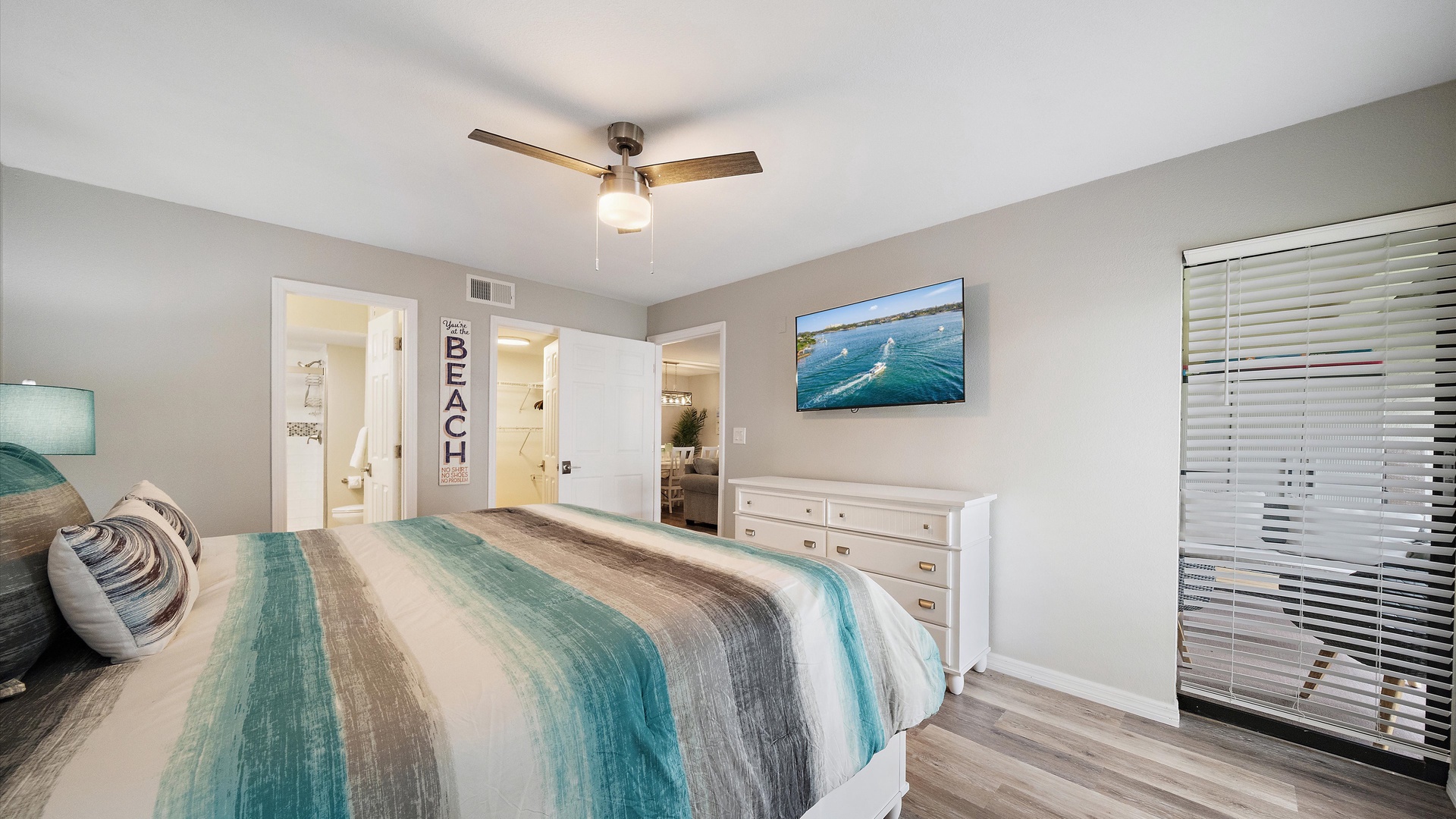 Your serene coastal primary bedroom features soothing aqua accents, a warm ceiling fan, and plenty of natural light streaming through the blinds—perfect for unwinding after a day by the water.