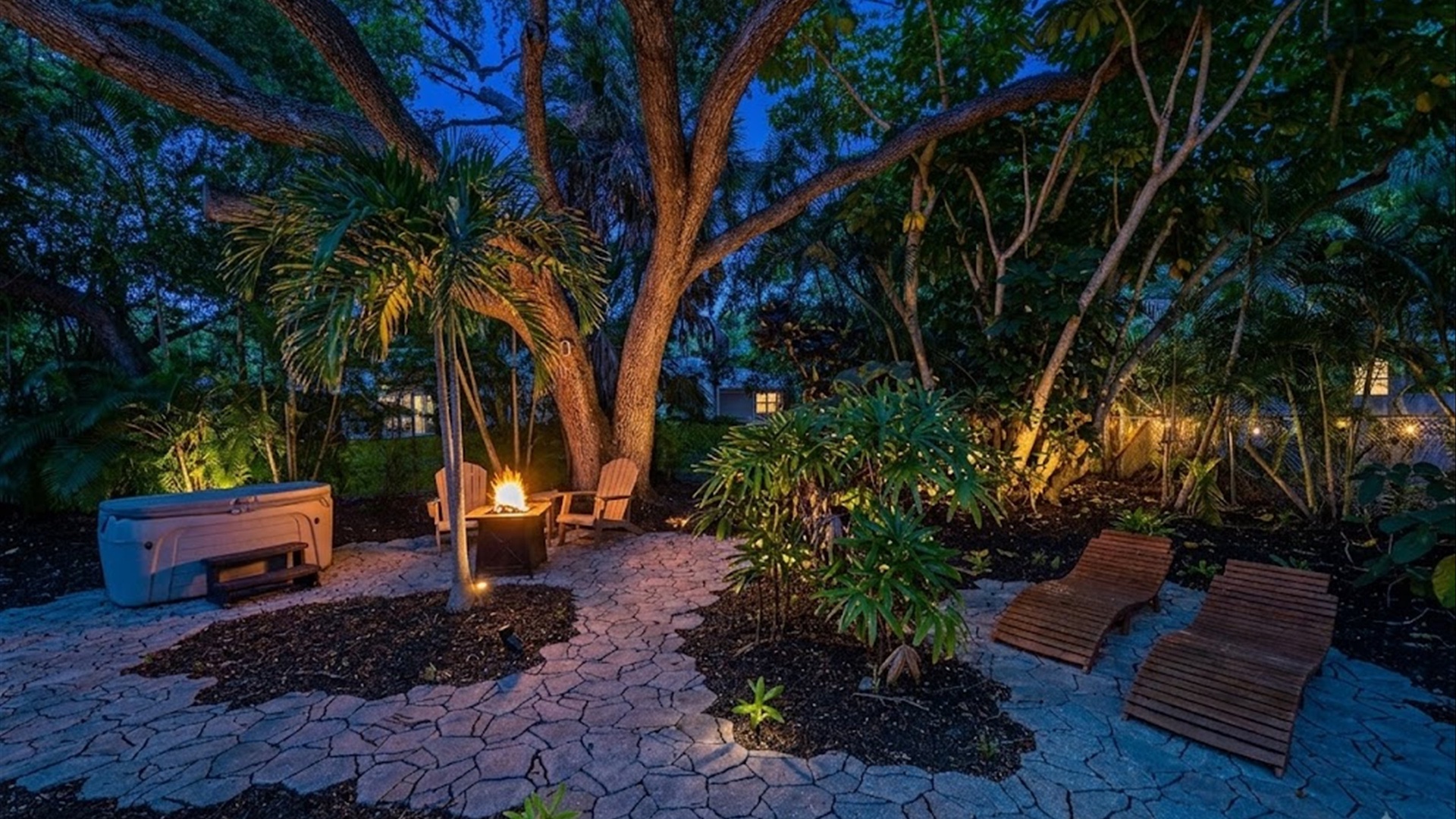 This tranquil evening retreat features a fire pit with seating, a private hot tub, and loungers nestled among lush tropical landscaping—perfect for unwinding under the stars.
