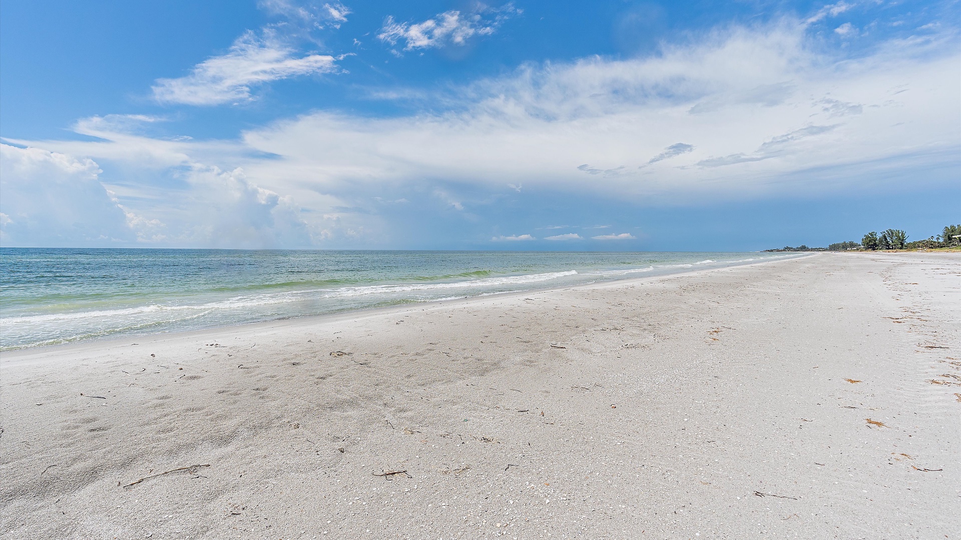 A wide sandy beach stretches along the calm sea under a blue sky with scattered clouds, with a distant shoreline dotted with trees.