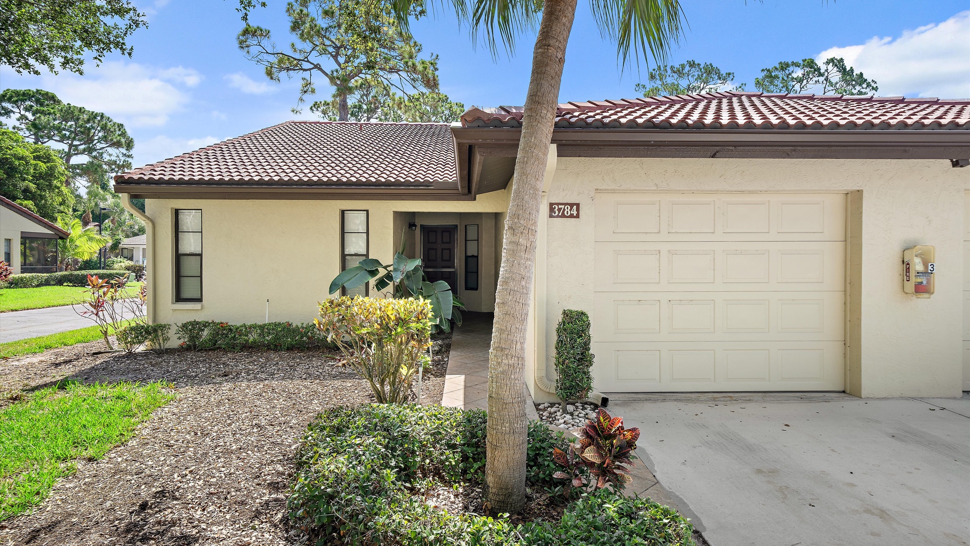 A charming single-story home with terracotta roof tiles, lush tropical landscaping, and palm trees, nestled in a peaceful residential neighborhood.