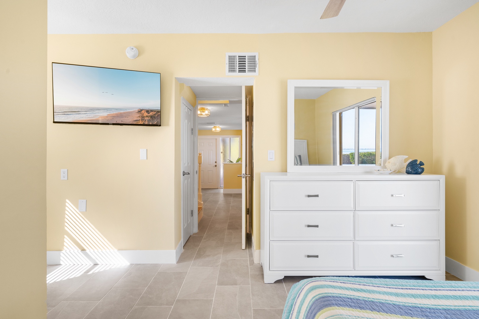 Your spacious bedroom offers coastal comfort with a white dresser, beachside artwork, and natural light streaming across the tiles.