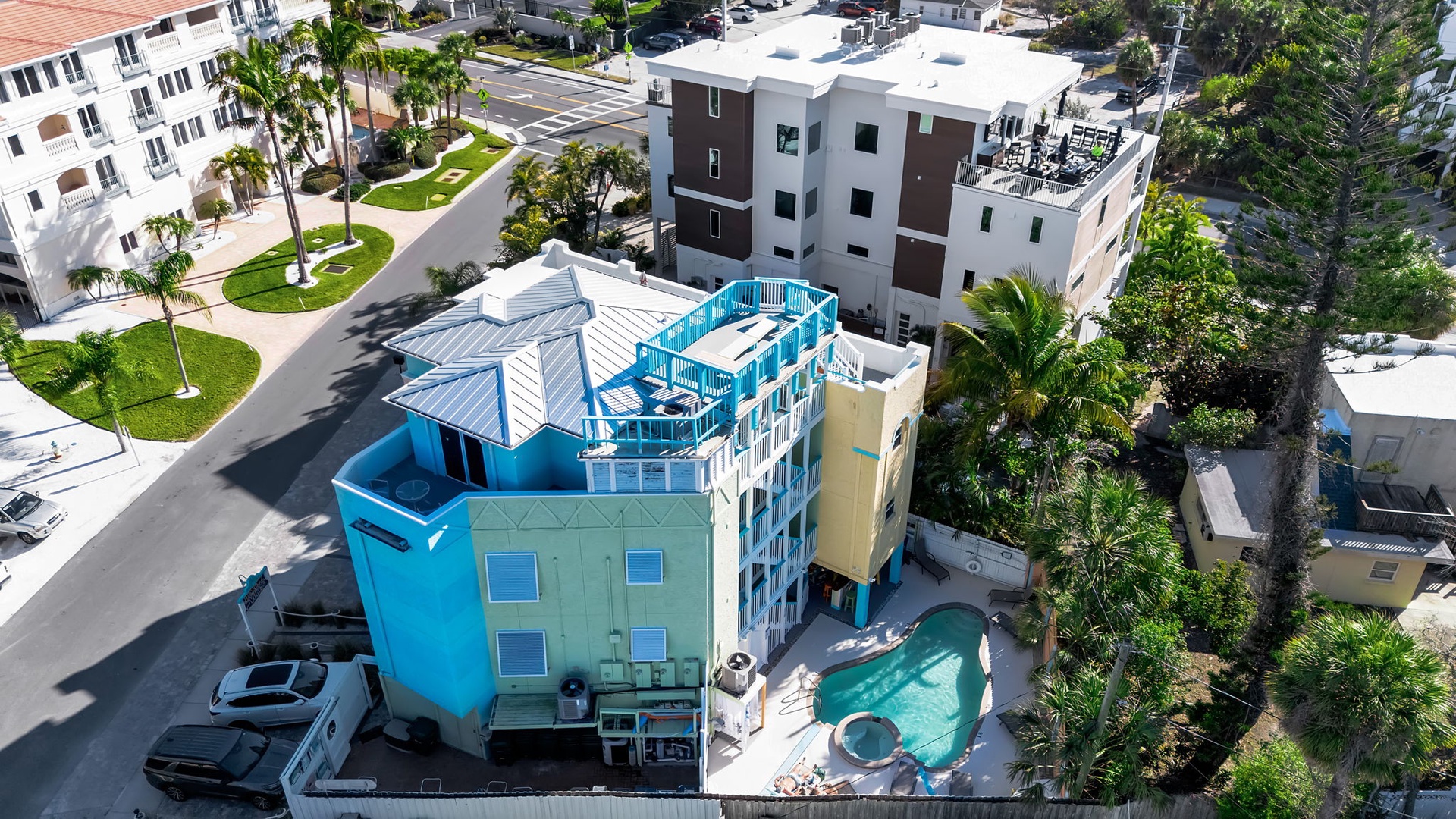 Aerial view of a modern vacation rental nestled in a vibrant neighborhood with palm trees, nearby residential buildings, and private pool area visible below.