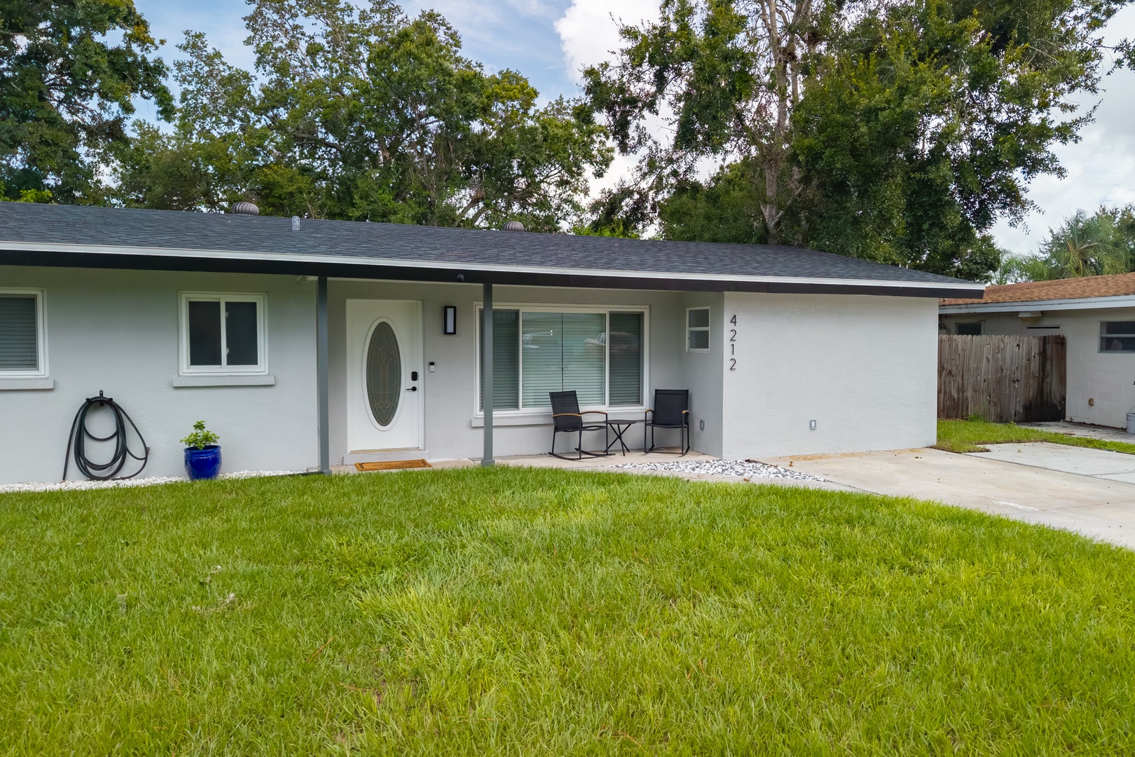 Modern single-story home with covered porch seating and private driveway, surrounded by lush mature trees in a quiet residential neighborhood.