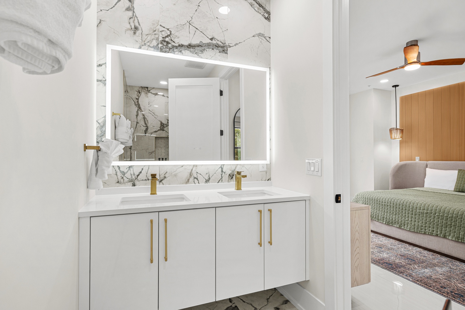 Step into your ensuite, where sleek marble walls frame dual vanities with gold hardware and LED-lit mirrors. The open door offers a glimpse of your cozy bedroom sanctuary just beyond.