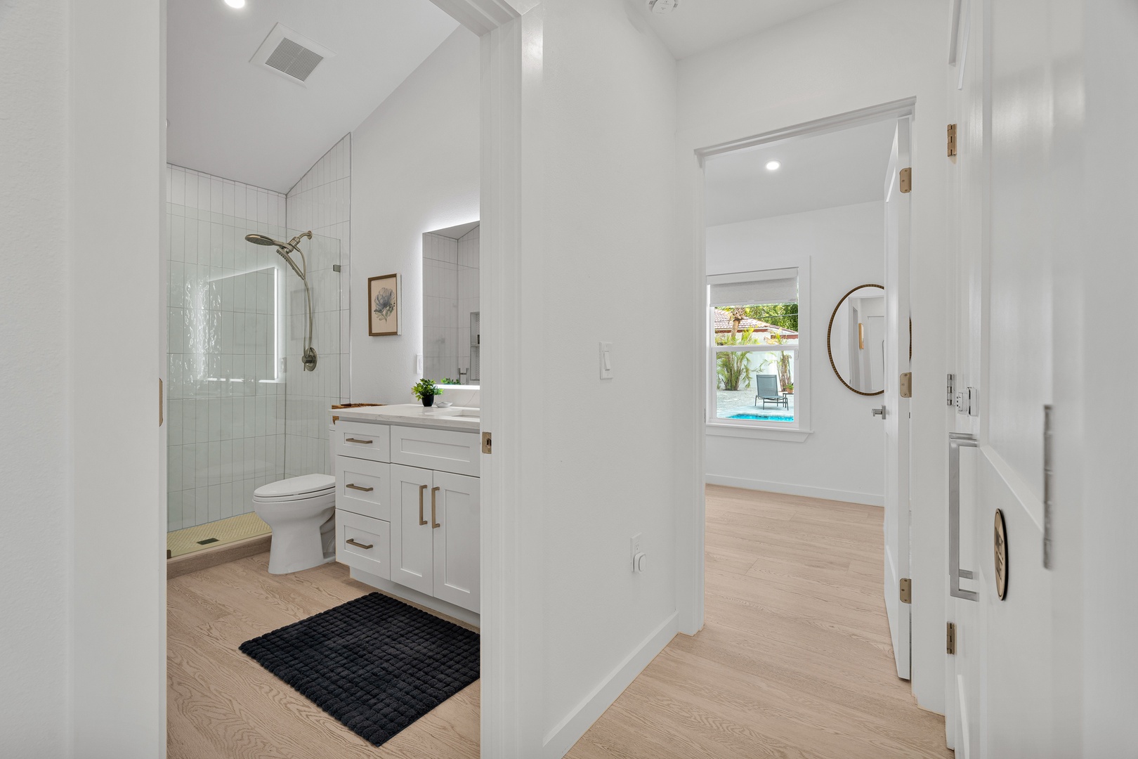 Your private bathroom gleams with bright, clean lines—walk-in shower, soft lighting, and a glimpse of the sparkling pool just beyond the hallway.