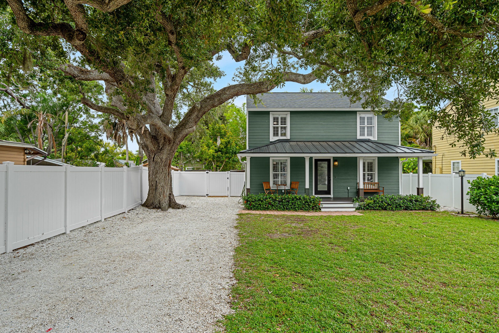 Charming two-story rental home with inviting porch seating, shaded by a grand oak tree, featuring a fenced yard and gravel driveway for easy parking.