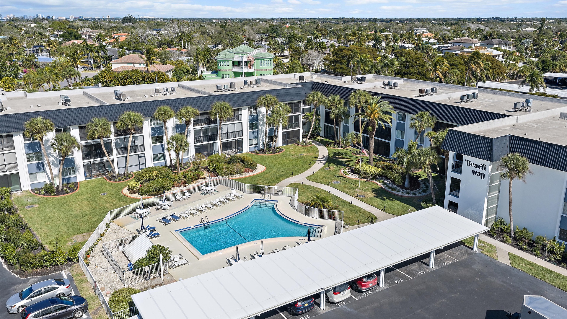 Aerial view of Beach Way resort featuring modern buildings, swimming pool, and tropical landscaping in a residential neighborhood.