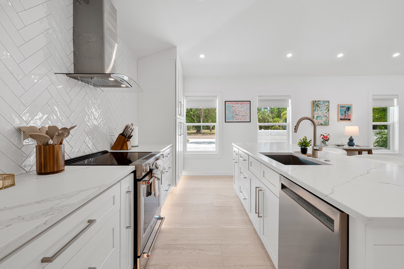 This bright, spacious kitchen invites you to prepare meals surrounded by gleaming white surfaces, modern appliances, and natural light streaming through large windows overlooking lush greenery.