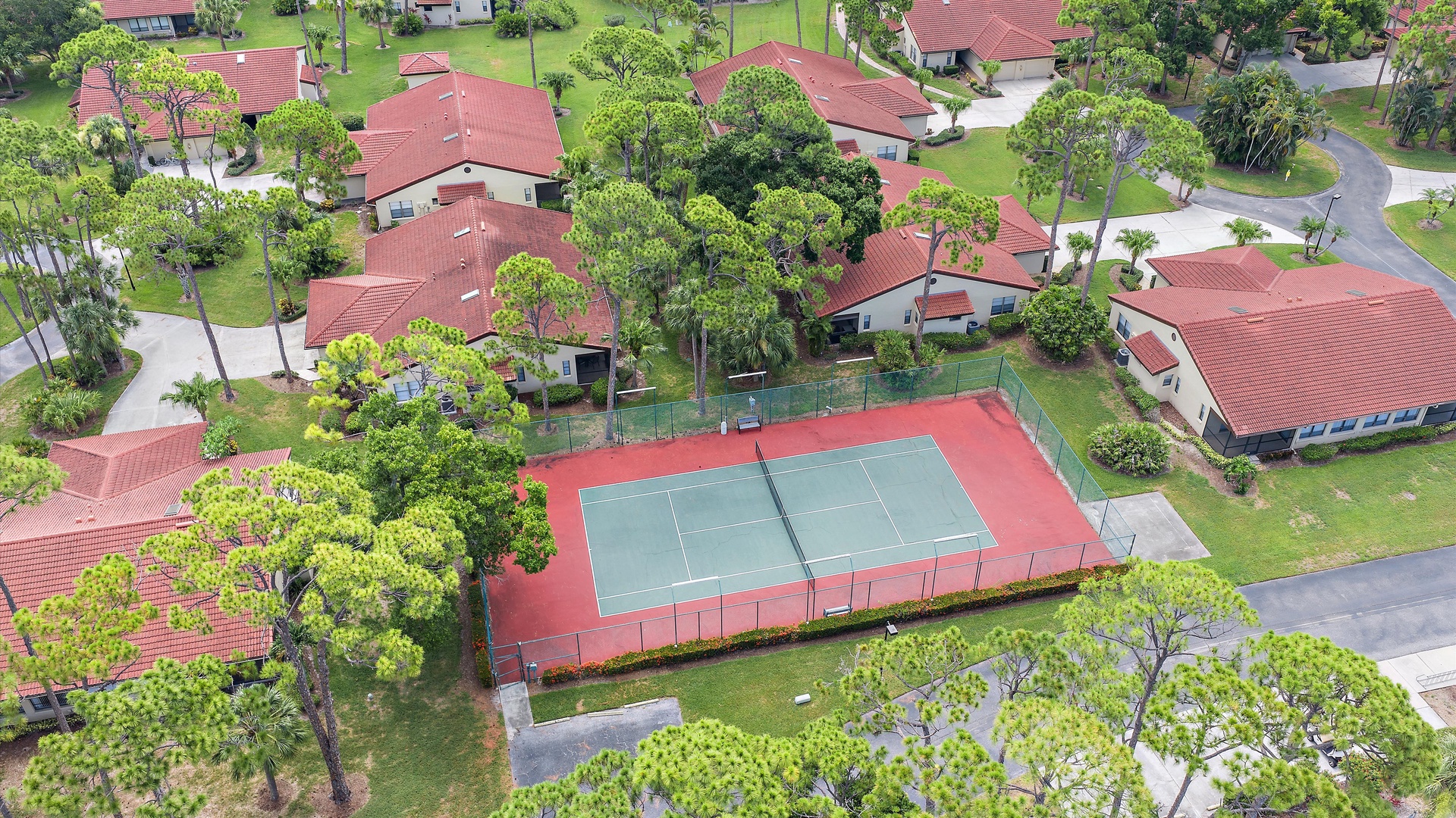 Aerial perspective of a residential community featuring a pickle ball court, red-tiled roof homes, and lush tropical landscaping with palm trees throughout the neighborhood.