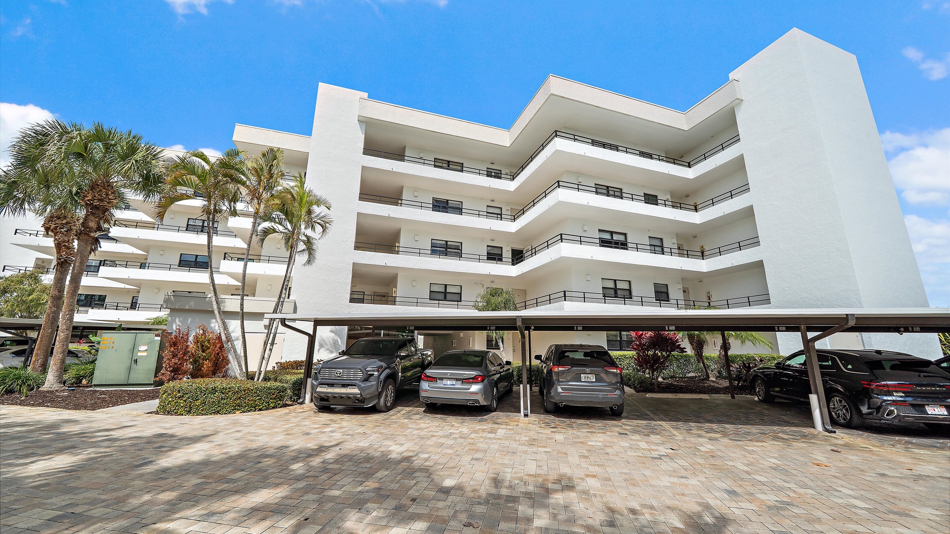 Modern white building with multiple balconied floors and covered parking area surrounded by tropical palm landscaping.