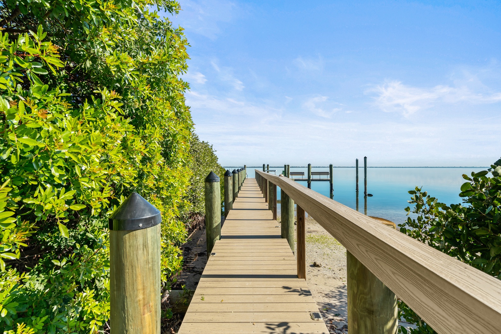 Wooden boardwalk leads through lush tropical vegetation to calm waters, offering peaceful waterfront access.