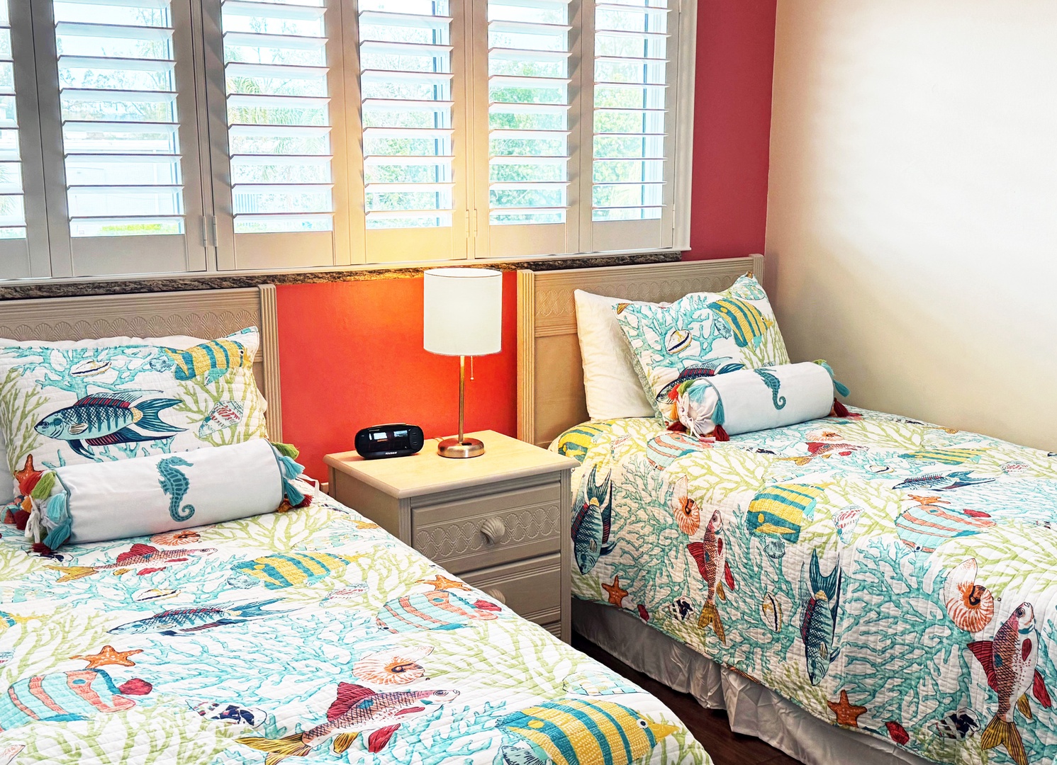 Wake up in this bright twin bedroom where colorful fish-themed bedding meets natural shuttered windows and warm coral accents.