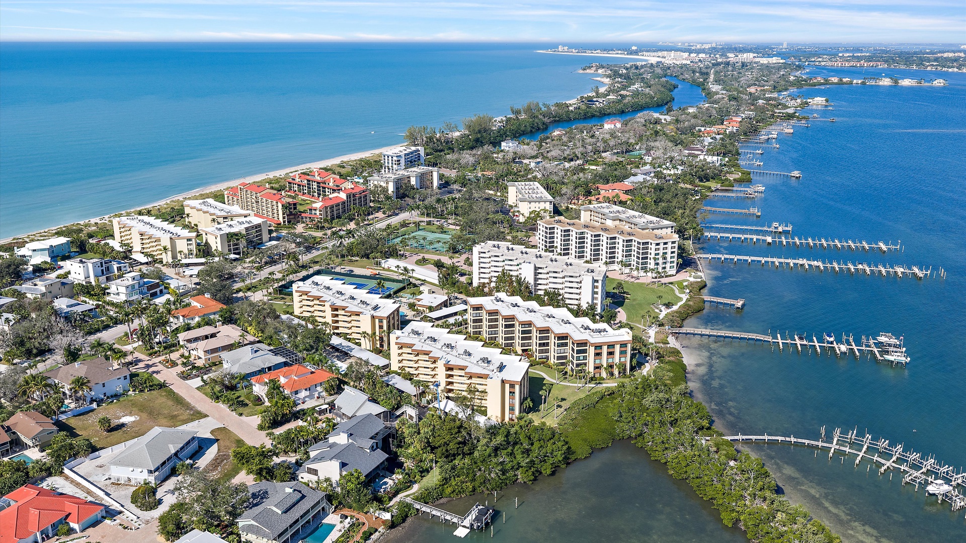 Aerial view of coastal area featuring beachfront buildings, marina with boat docks, and residential neighborhoods near pristine blue waters.