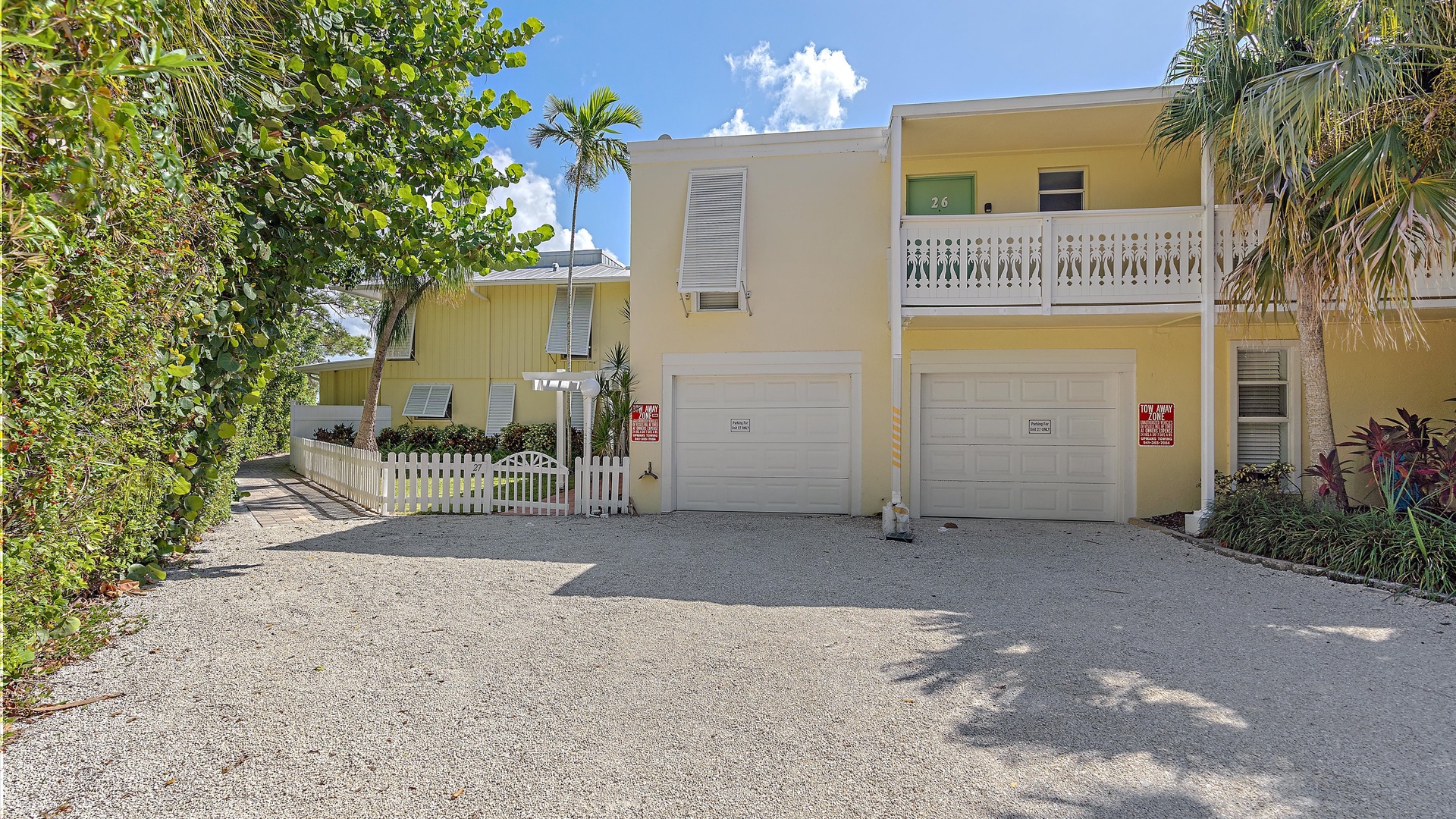 Two-story yellow building with a covered balcony, white shutters, and two closed white garage doors. Tropical plants and trees surround the gravel driveway in the foreground.
