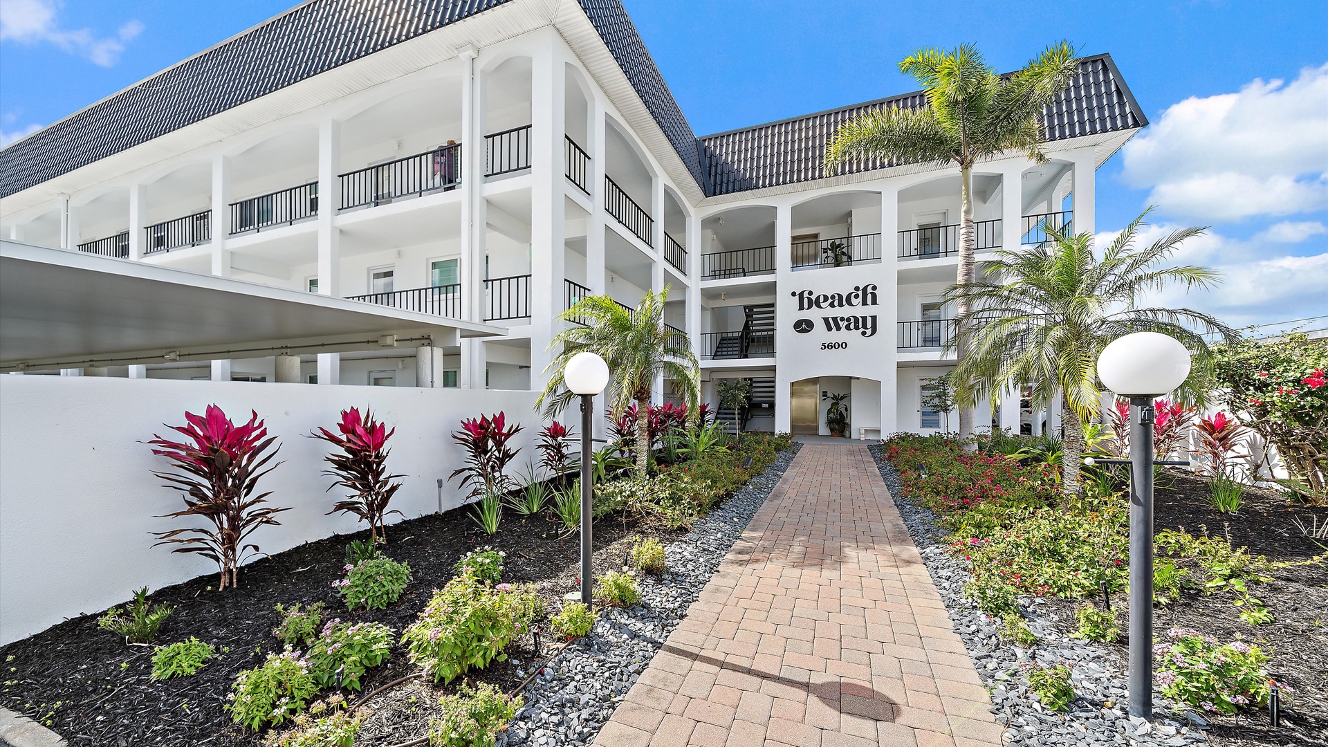 The Beach Way property welcomes guests with tropical landscaping, palm trees, and a charming brick pathway leading to the main entrance.