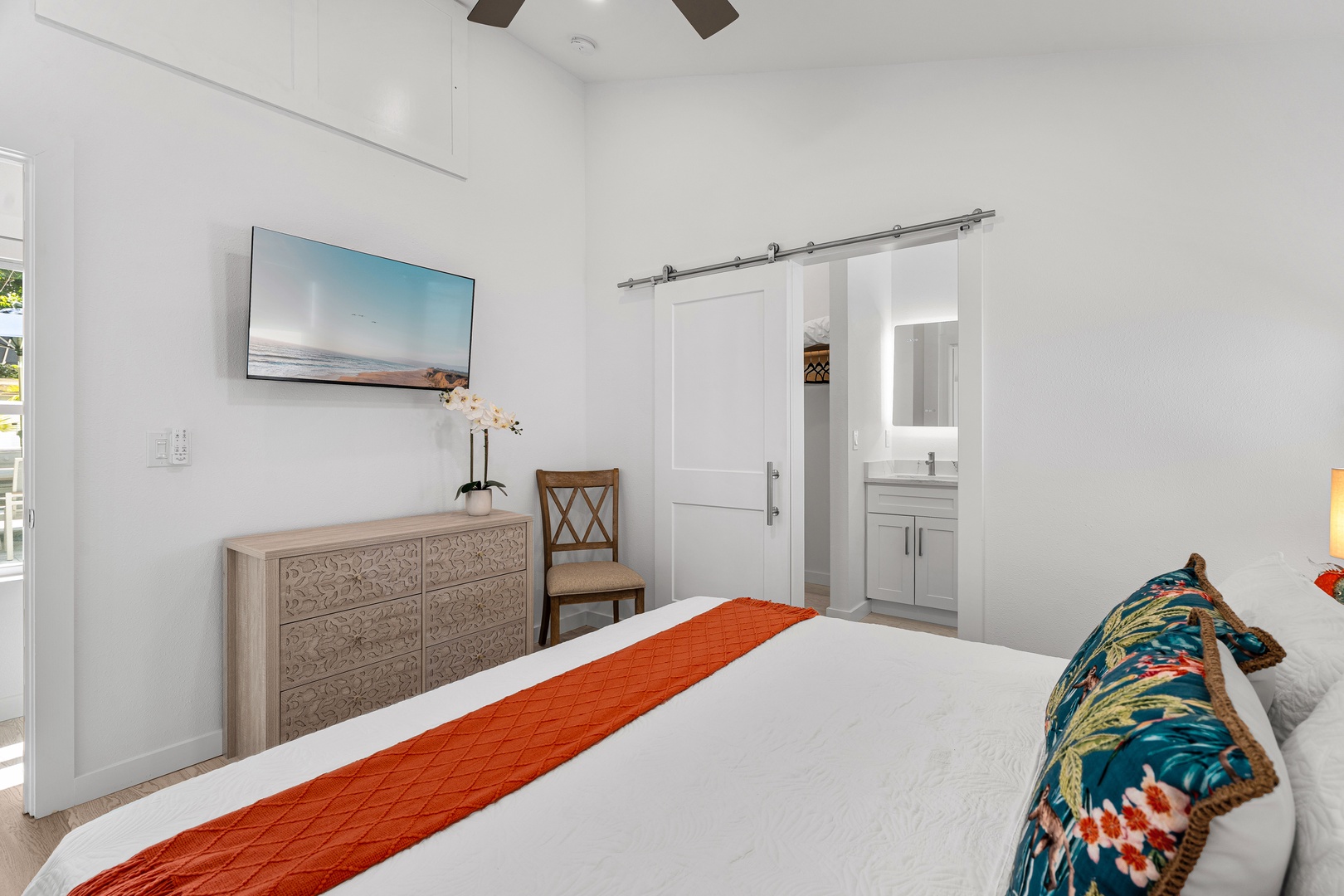 Your serene bedroom features crisp linens, vaulted ceilings with a ceiling fan, and a wall-mounted TV for restful evenings. A charming barn door slides open to reveal your private ensuite bathroom.