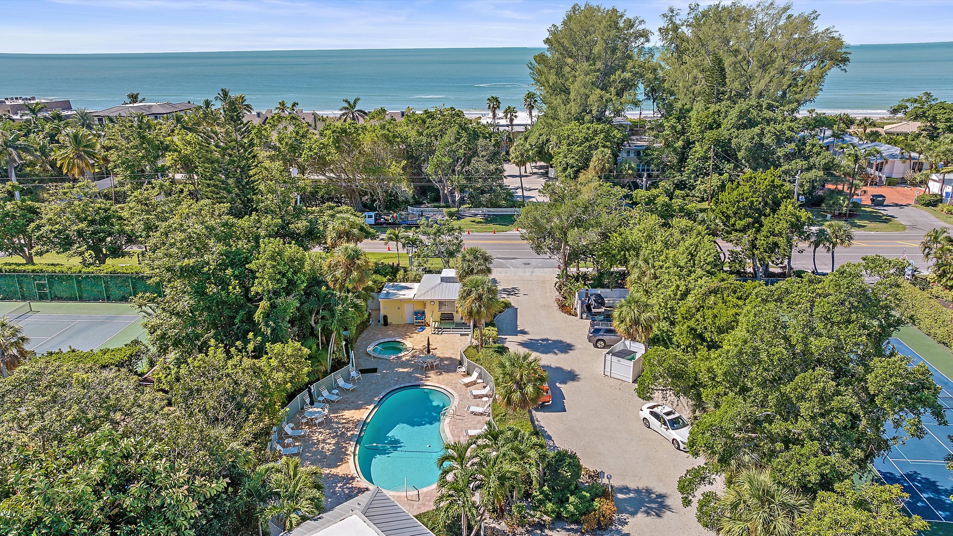 Aerial view of a coastal area featuring a swimming pool, tennis court, parking lot, and trees, with the ocean and beach in the background.