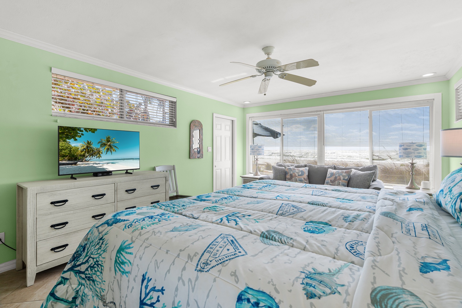 Your bright coastal bedroom features tropical bedding, a large TV, and stunning ocean views through expansive windows.