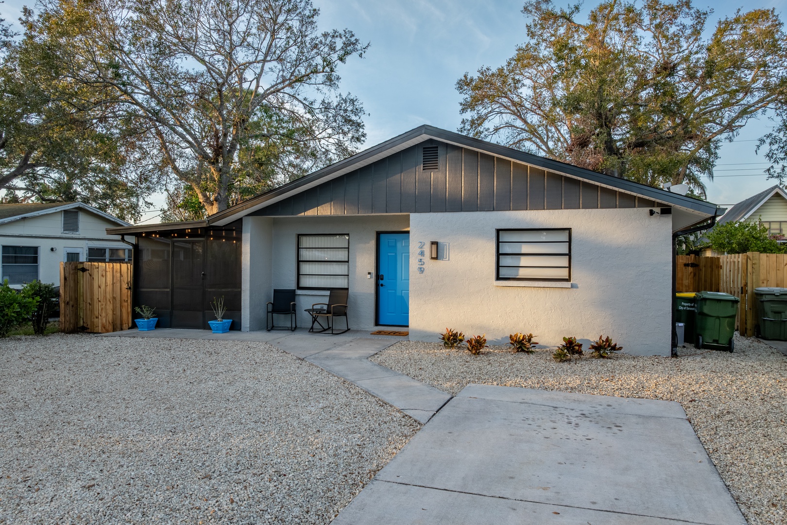 Modern single-story home with bright blue door and covered porch seating area, surrounded by mature trees in quiet residential neighborhood.