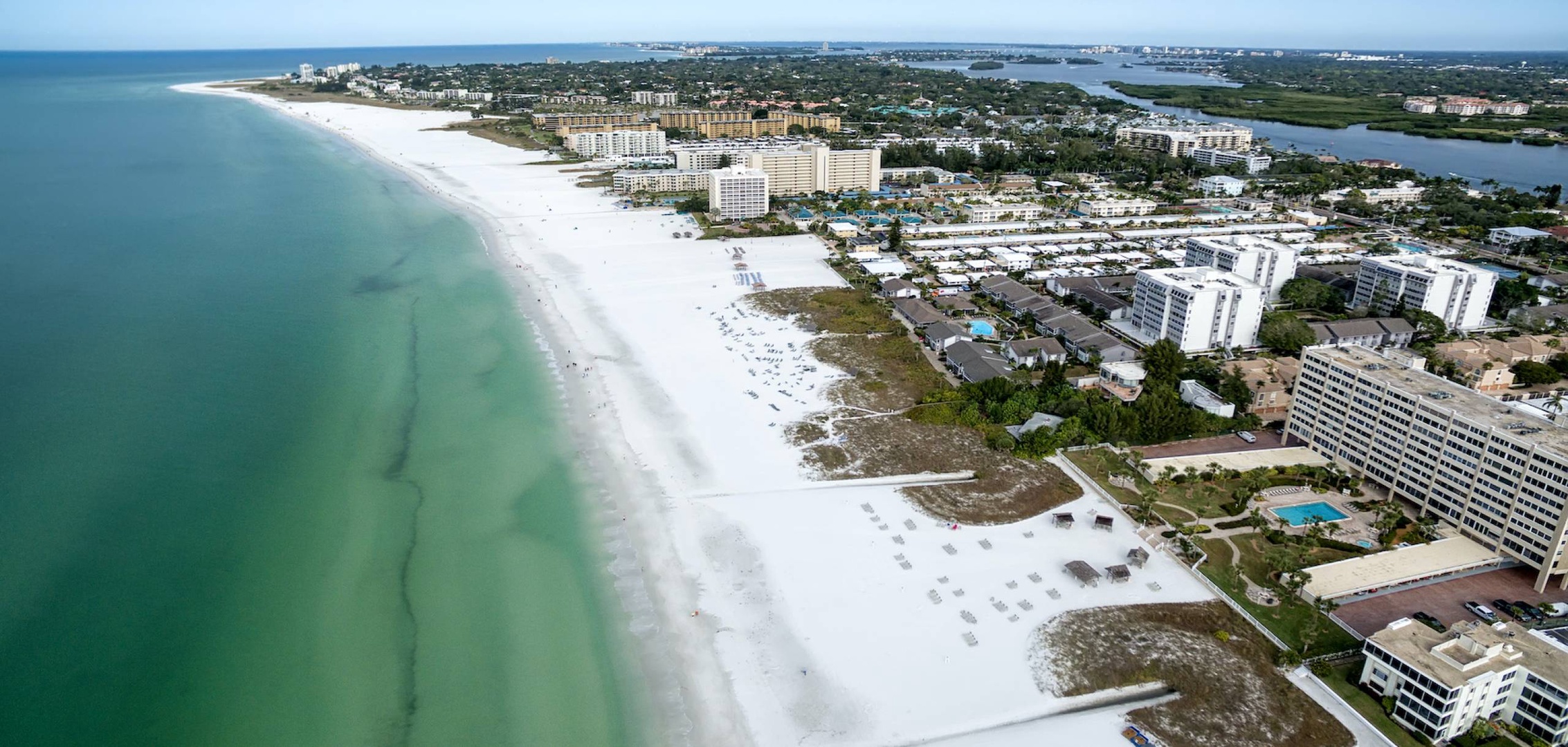 Aerial view showcasing the pristine white-sand beaches and turquoise waters of this coastal area, with nearby properties and scenic landscapes stretching into the horizon.