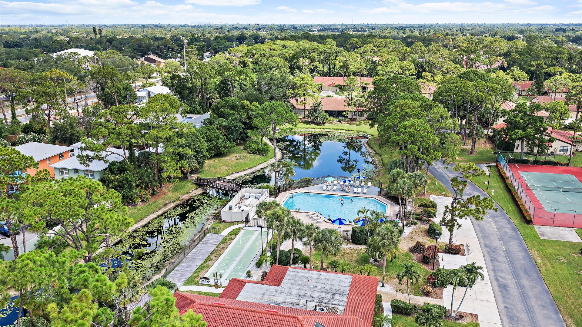 Aerial view of a lush residential community featuring a sparkling pool, tennis court, and tranquil lake surrounded by mature trees and walking paths.