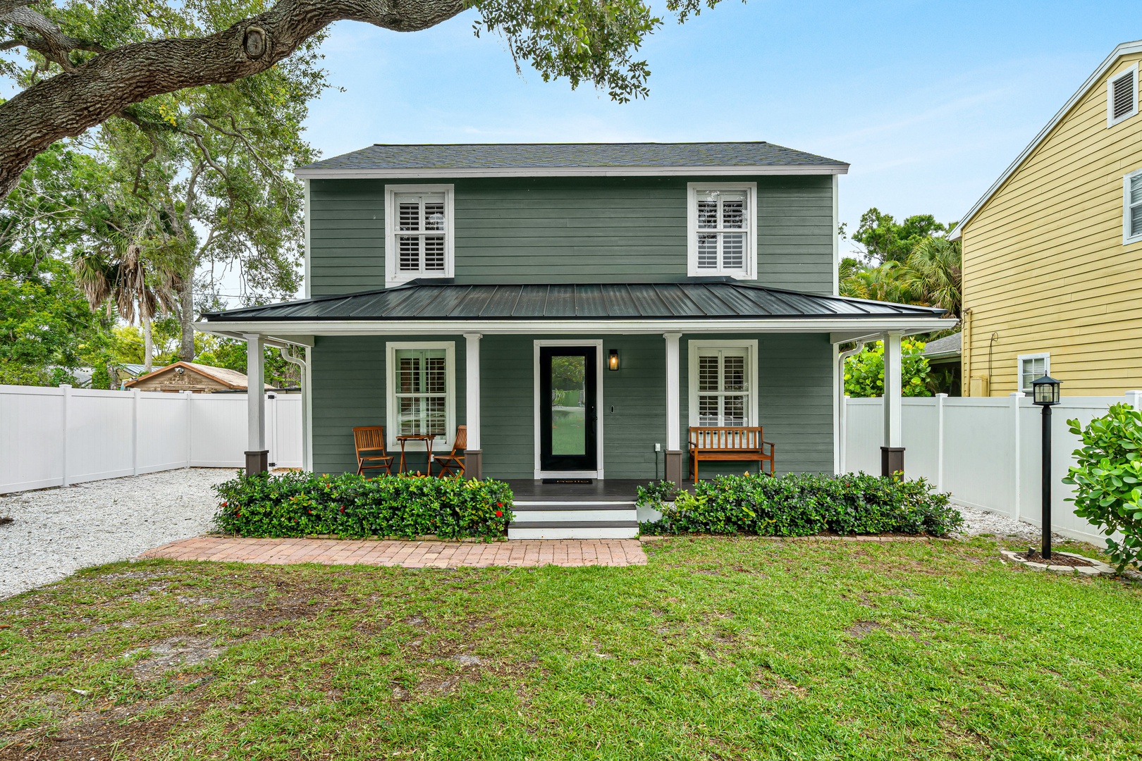 A charming two-story home with classic coastal styling, featuring a welcoming covered porch with rocking chairs, manicured hedges, and mature shade trees. The property offers a peaceful residential setting with privacy fencing and easy front-yard parking.